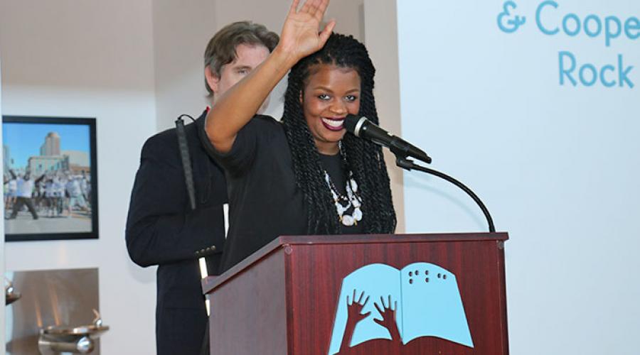 a woman stands behind a podium waving at the audience