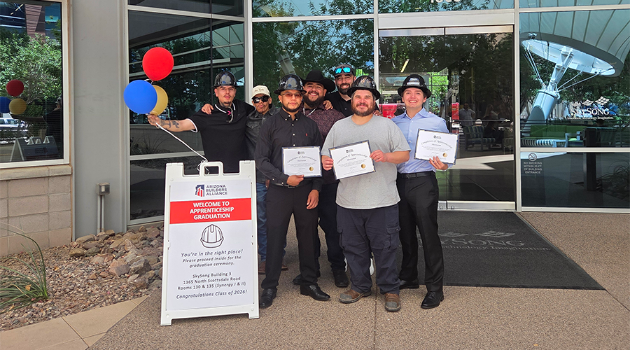 A group of people stands outside a building, smiling and holding certificates for an apprenticeship graduation.