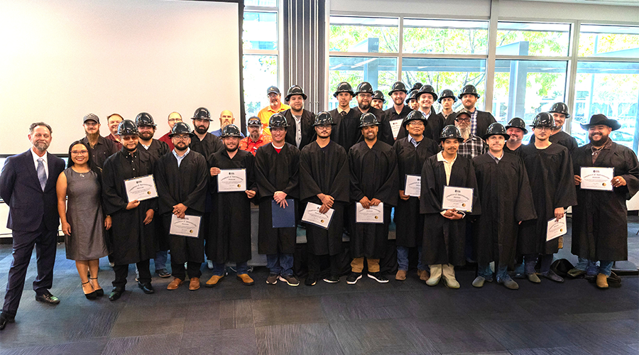 A group of graduates in black gowns and hard hats, holding certificates, poses for a photo in a bright indoor setting.