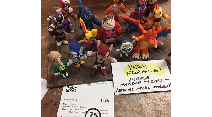various colorful clay figurines stand on a tray; a label reads "Very fragile, please handle with care, special needs student"; another label reads "third place"