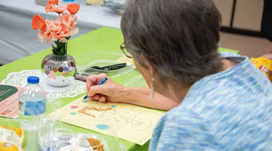 An older woman colors a drawing at a table with flowers and snacks.