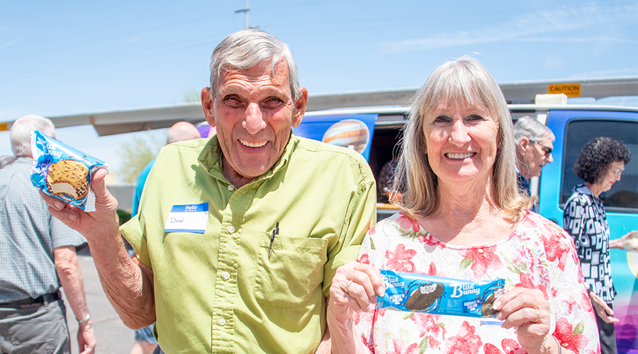 Two people smiling and holding ice cream sandwiches in front of a blue van on a sunny day.