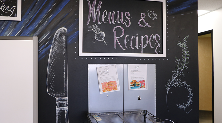 Two menus are tacked onto a display wall that replicates a black board.  The wall features a chalk-like drawing of a knife and a sign that reads “Menus & Recipes.”