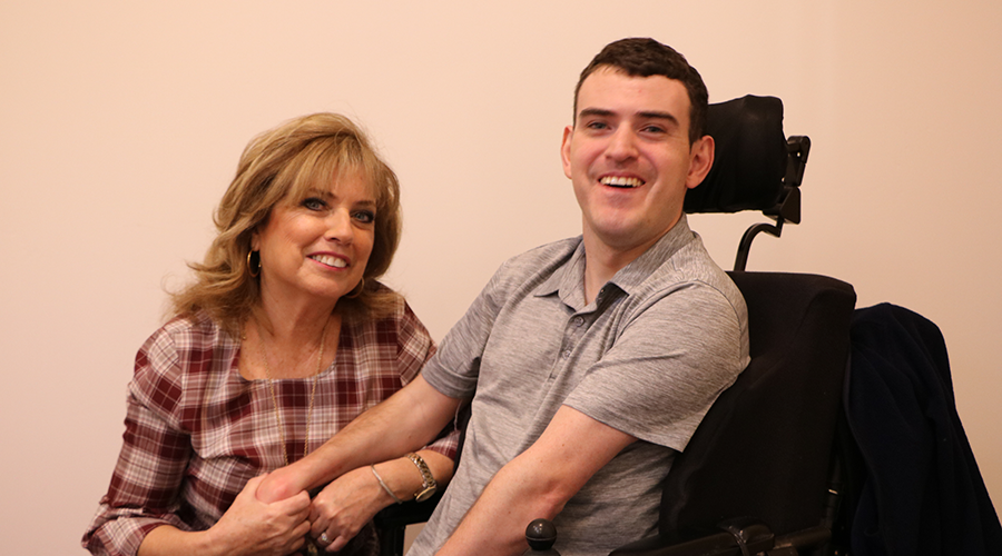 a woman in a plaid blouse sits next to a 20-year old man who is in a wheel chair