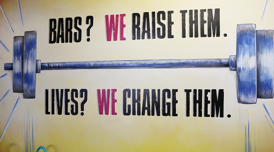 A graphic image of barbells is painted onto a yellow wall.  Above the drawing are the words, “Bars?  We raise them.”  Below the drawing are the words, “Lives?  We change them.”