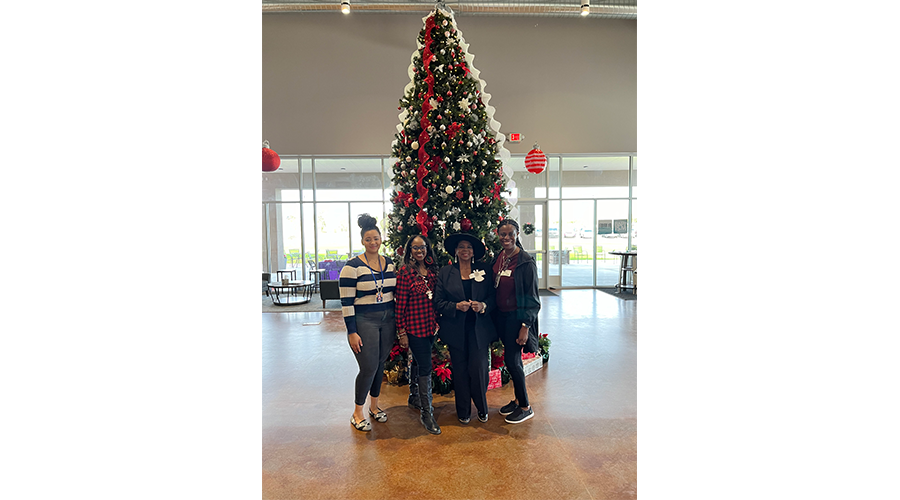 Four people standing in front of a large, decorated Christmas tree indoors.