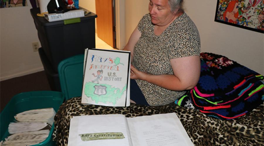 A woman holds up a handmade book with a drawing of cartoon figure and a map of the United States on the cover; the words "Ray's America - US History" display at the top