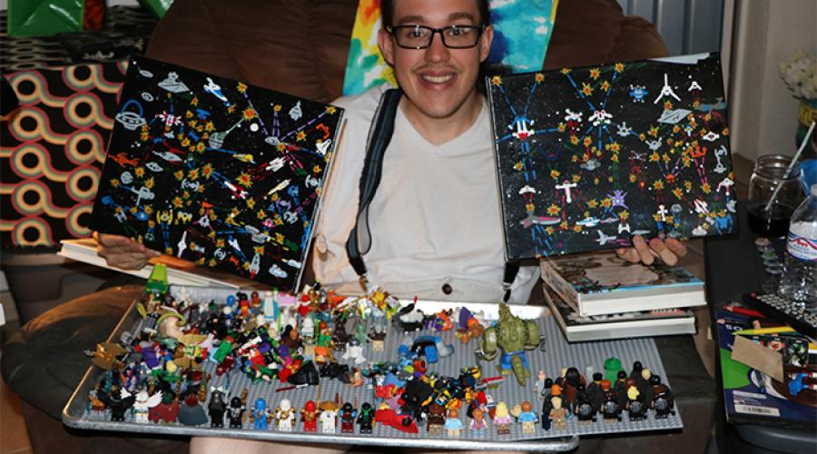 A young man sits on a sofa, holding up two paintings; a tray holding various Lego creations rests on his legs.