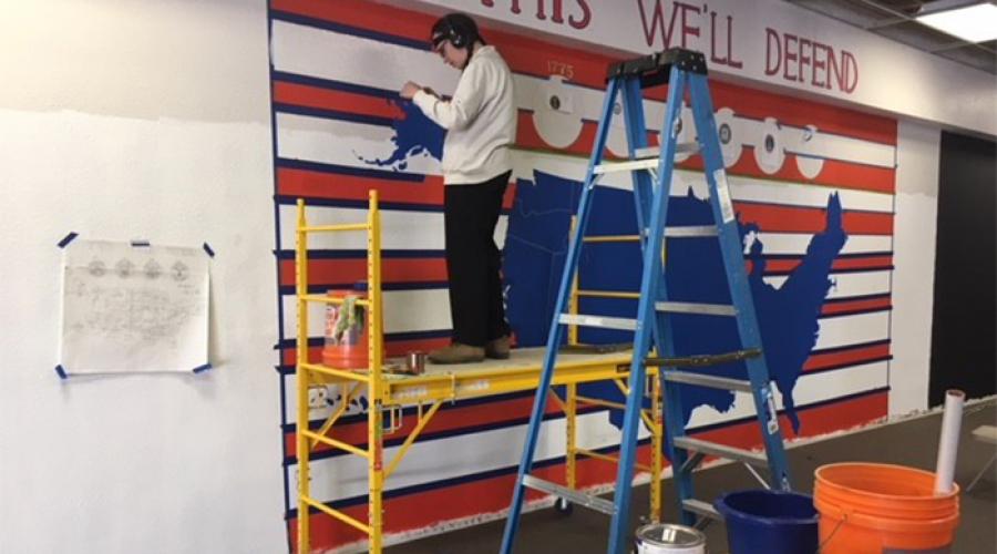 Man standing on scaffold paints mural that includes a map of the United States; the words "This we'll defend" display at the top