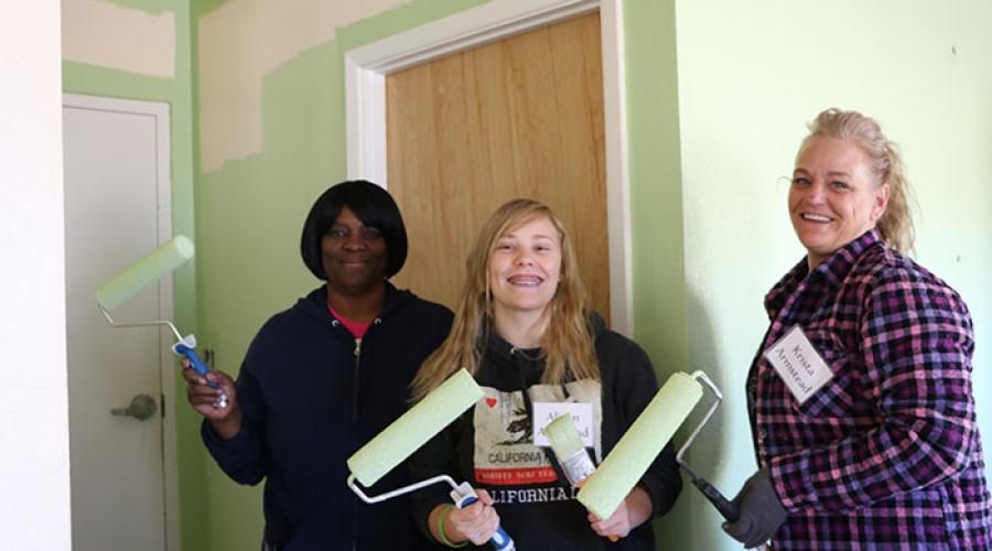 Three women holding paint rollers pose for pictures inside of a home