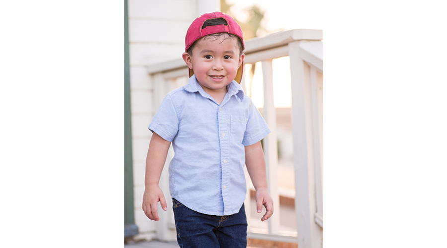 Young boy wearing a red baseball cap turned backwards, stands on a porch and smiles at the camera.