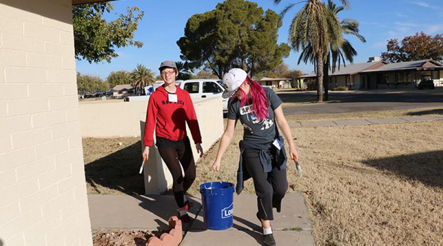 Two women walking along the outside of a home carrying painting bucket and painting supplies