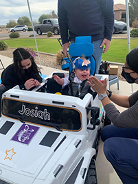 woman applauds as a toddler sits behind the wheel of toy car, laughing