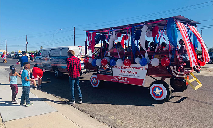 A group of adults sit inside a full-sized wagon with canopy. The wagon is decorated with red, white and blue streamers, stars and balloons.