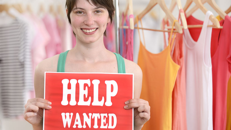 a woman stands inside a clothing store holding a sign that reads "Help Wanted"