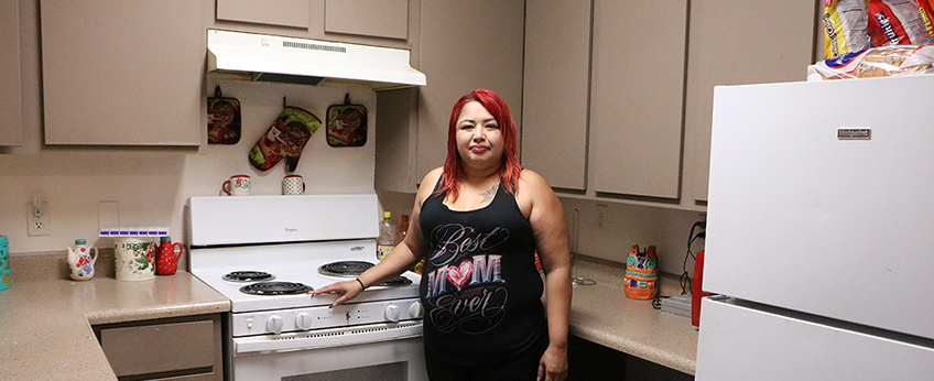 A woman wearing a “best mom ever” tank top and shorts stands next to the stove in a kitchen